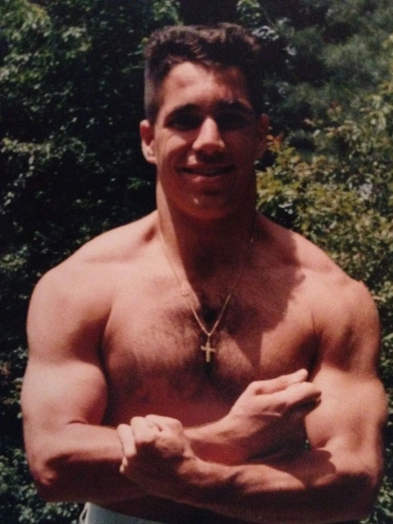 Franco Zuccoli as a young man, shirtless with his arms crossed, smiling outdoors in front of trees